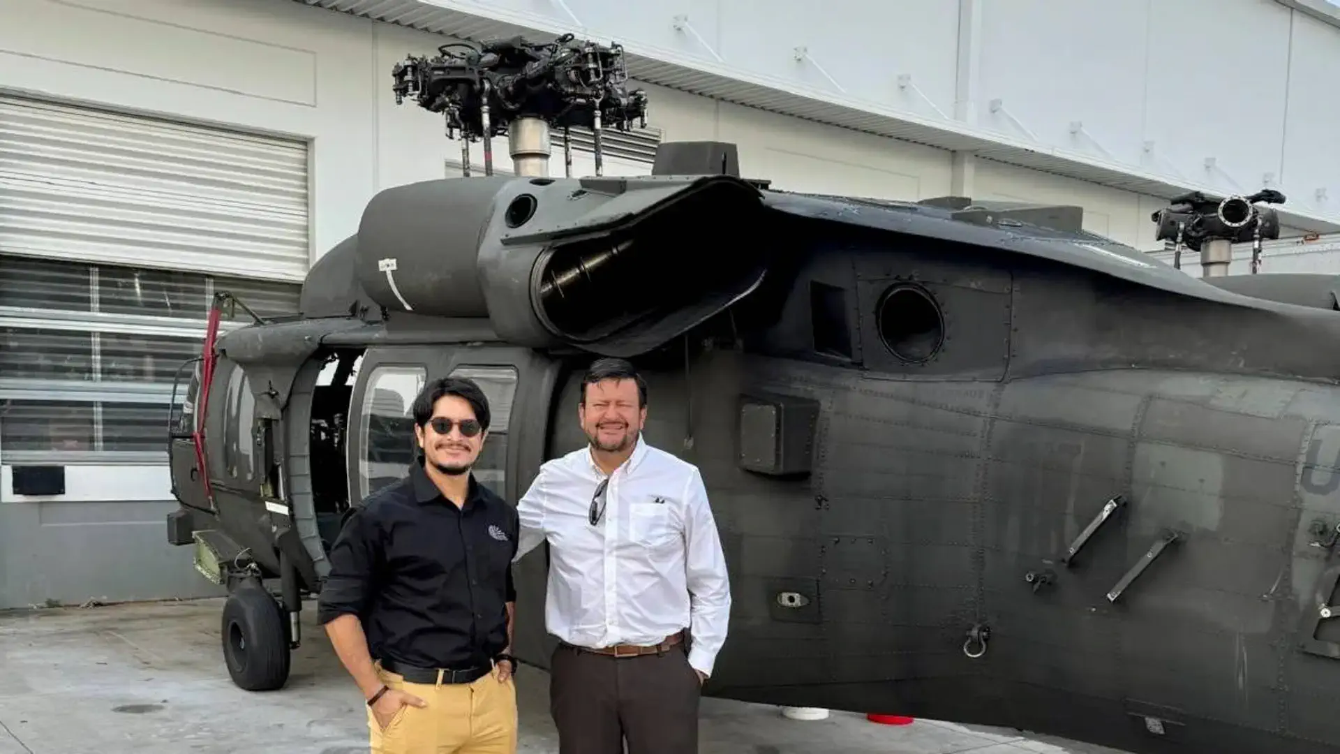 Two men standing in front of a military helicopter parked outside a building.